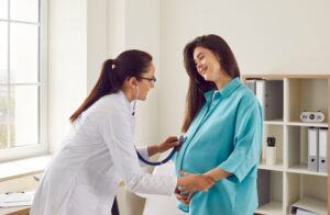 Doctor obstetrician with stethoscope examines happy young pregnant woman patient